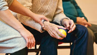 A person helps an elderly person holding a stress ball.  Image by Matthias Zomer via Pexels.